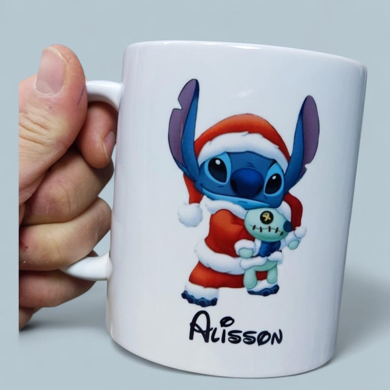 K&ouml;nnte beinhalten: Wei&szlig;e Keramiktasse mit einem blauen Cartoon-Charakter in einem Weihnachtsmann-Outfit, der ein kleines Pl&uuml;schtier h&auml;lt. Auf der Tasse steht der Name "Alisson" in schwarzer Schrift. Die Tasse hat einen wei&szlig;en Henkel.
