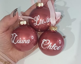 Könnte beinhalten: Drei bordeauxfarbene Weihnachtskugeln mit goldenen Kappen und rosa Bändern. Jede Kugel trägt einen Namen in weißer Schrift: "Lee-an", "Louina" und "Chloe". Die Kugeln sind rund und glänzend.