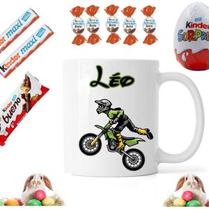 Peut inclure: Une tasse en céramique blanche avec un motif de motard de motocross vert et noir et le nom "Léo" imprimé dessus. La tasse est entourée de divers articles de chocolat et de bonbons, notamment des barres de chocolat Kinder, des œufs Kinder Surprise et des bonbons au chocolat Schoko Bons.