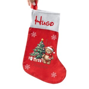 May include: Red Christmas stocking with a white cuff, personalized with the name "Hugo" in red script. The stocking features a Christmas tree, a teddy bear wearing a Santa hat and scarf, and the text "1st Christmas".