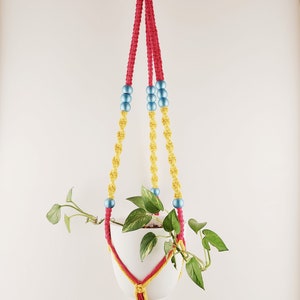 May include: A macrame plant hanger with three strands of red cord and yellow cord with blue beads. A white pot with a green plant is hanging from the macrame.