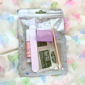 May include: A clear plastic bag containing nail care items. Includes a pink nail file, a small bottle of nail glue, a purple sanding block, a wooden cuticle stick, and an alcohol prep pad. The bag has a heart and butterfly pattern.