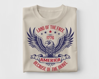 Puede incluir: Una camiseta beige doblada con un diseño patriótico. El diseño incluye una ilustración detallada de un águila con las alas extendidas, con el texto "LAND OF THE FREE SINCE 1776 AMERICA BECAUSE OF THE BRAVE" en rojo y azul.