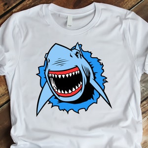 May include: White t-shirt with a blue and red cartoon shark graphic. The shark is shown with its mouth open and teeth visible, as if it is about to bite.