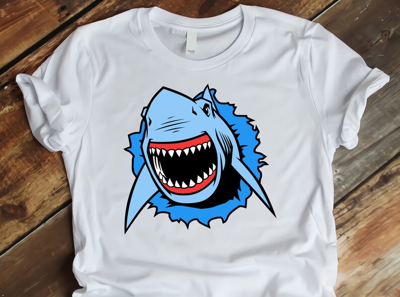 May include: White t-shirt with a blue and red cartoon shark graphic. The shark is shown with its mouth open and teeth visible, as if it is about to bite.