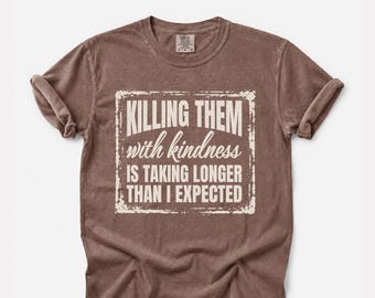 Puede incluir: Una camiseta marrón de manga corta con un gráfico cuadrado blanco desgastado en el pecho. El texto dice: KILLING THEM with kindness IS TAKING LONGER THAN I EXPECTED. La camiseta presenta un cuello redondo y un acabado de tela lavada.