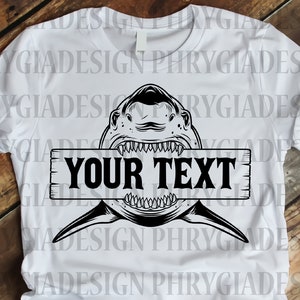 以下が含まれることがあります： 白いTシャツに、口を開けたサメの頭部の白黒イラストが描かれています。サメは「YOUR TEXT」と書かれた木製の看板を持っています。