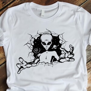 May include: White t-shirt with a black and white graphic print of an alien reaching out from a cracked wall.