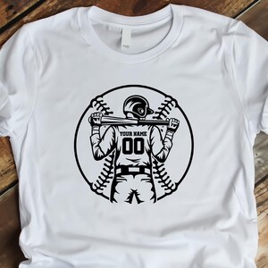 Op de afbeelding: Wit T-shirt met een zwart-witte afbeelding van een honkballer die een knuppel vasthoudt. De afbeelding bevat de tekst "YOUR NAME" en het nummer "00".