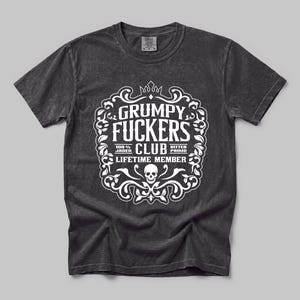 May include: A charcoal grey short-sleeve t-shirt featuring a white graphic print. The design includes a decorative frame with a crown, a skull, and the text: GRUMPY FUCKERS CLUB, 100% JADED, BITTER PROUD, and LIFETIME MEMBER in a bold, stylised font.