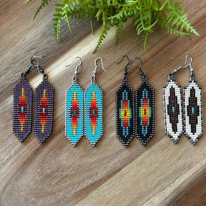 May include: Five pairs of beaded earrings in various colors, including purple, turquoise, black, and white. Each pair features a rectangular design with a central pattern of red, yellow, and blue. The earrings are displayed on a wooden surface.