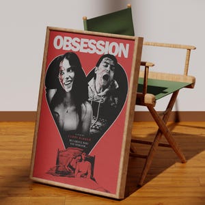 Puede incluir: Póster de película enmarcado con el título "OBSESSION" en blanco. El póster presenta un diseño en forma de corazón rojo con dos imágenes en blanco y negro de personas. Se apoya en una silla de director de madera.
