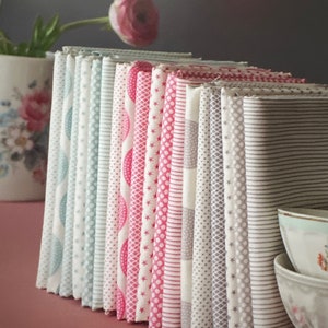 May include: A stack of folded fabric bundles in various patterns and colors, including light blue, pink, and gray. The fabrics feature designs such as polka dots, stars, and stripes. A vase with a pink flower is in the background.
