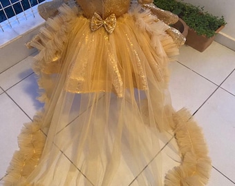 May include: A gold sequined dress with a tulle skirt and a long tulle train. The dress has a bow on the back and is decorated with ruffles.