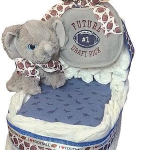 Puede incluir: Un pastel de pañales para bebé en forma de cuna, decorado con cintas de temática futbolística y un elefante de peluche gris. El pastel está coronado con una almohada gris y blanca con el texto "Future #1 Draft Pick".