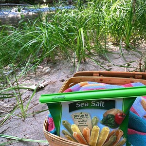Puede incluir: Una bolsa verde de patatas fritas con sal marina con un clip verde. La bolsa dice "50% menos de grasa que las patatas fritas l&iacute;deres" y "Sin sabores artificiales ni conservantes". La bolsa est&aacute; en una cesta de picnic de mimbre con una manta de colores.