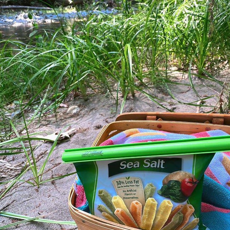 Puede incluir: Una bolsa verde de patatas fritas con sal marina con un clip verde. La bolsa dice "50% menos de grasa que las patatas fritas l&iacute;deres" y "Sin sabores artificiales ni conservantes". La bolsa est&aacute; en una cesta de picnic de mimbre con una manta de colores.