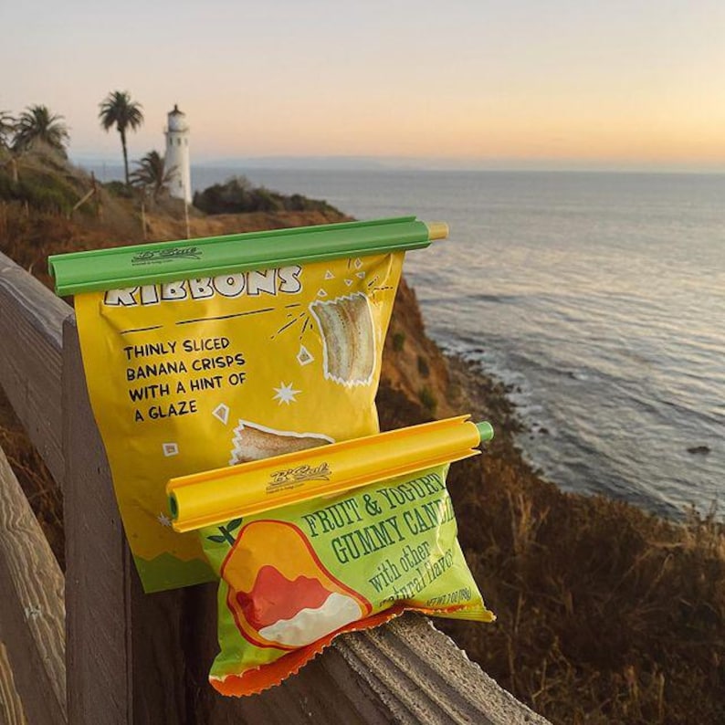 Puede incluir: Dos clips de almacenamiento de alimentos reutilizables de color amarillo y verde con un dise&ntilde;o amarillo y verde. El clip superior tiene el texto "Ribbons" y "Thinly sliced banana crisps with a hint of a glaze". El clip inferior tiene el texto "Fruit & Yogurt Gummy Candy" y "with other natural flavors".