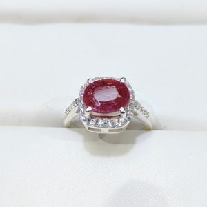 Könnte beinhalten: Ein silberner Ring mit einem großen, ovalen Rubin, der von einem Heiligenschein aus kleineren, weißen Edelsteinen umgeben ist.