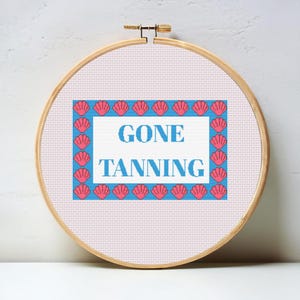 Könnte beinhalten: Ein Kreuzstich-Stickrahmen mit den Worten "GONE TANNING" in Blau, umgeben von einem blauen Rand und rosa Muschelmotiven. Der Rahmen ist vor einem weißen Hintergrund platziert.