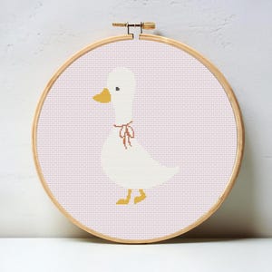 Peut inclure: Broderie au point de croix d'un canard blanc avec un bec et des pattes jaunes, portant un ruban rose. Le canard est centré sur un fond rose clair, encadré par un cercle à broder en bois.