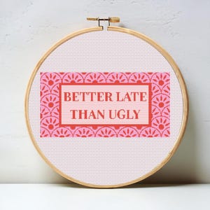 May include: A pink and red cross-stitch embroidery hoop with the saying "Better Late Than Ugly" in red lettering.