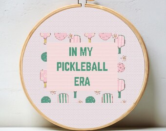 May include: A pink and green cross-stitch embroidery hoop with the text "In My Pickleball Era" surrounded by a pattern of pink paddles and green leaves.