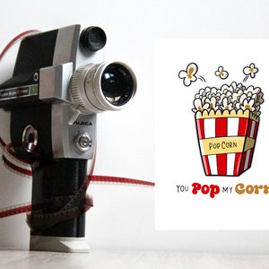 May include: A vintage Super 8 film camera with a red and white film reel wrapped around it. A white card with a cartoon image of a red and white striped popcorn bucket with popcorn spilling out and the text "You Pop My Corn".
