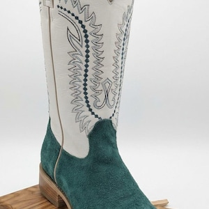 Op de afbeelding: Een hoge cowboylaars met een witte leren schacht voorzien van donkerblauw decoratief stiksel en een voet van blauwgroen suède. De laars heeft een vierkante neus, een gestapelde houten hak en een lichte zool met een opvallend contrast.