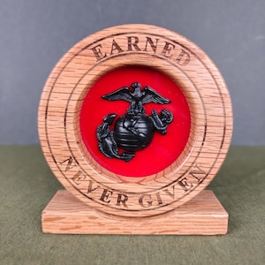 May include: A circular wooden display stand with a light brown wood grain finish. The frame features the engraved text EARNED NEVER GIVEN. Centered inside is a bright red circular background with a black United States Marine Corps emblem.