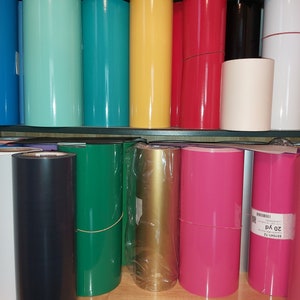 Könnte beinhalten: Ein Regal mit Rollen Vinyl in verschiedenen Farben, darunter Türkis, Gelb, Rot, Weiß, Schwarz, Grün, Gold, Rosa und Lila. Die Rollen befinden sich auf einem Holzregal mit schwarzem Metallrahmen.