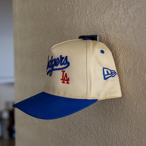 May include: A cream-colored baseball cap with a blue brim. The front features the blue embroidered text Dodgers and a red LA logo. A blue New Era logo is embroidered on the side panel. The cap is mounted on a black wall hook.