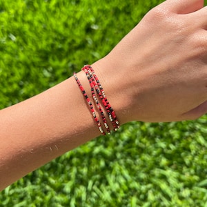 May include: A set of four beaded bracelets in red, black, and silver. The bracelets are stacked on a person's wrist.