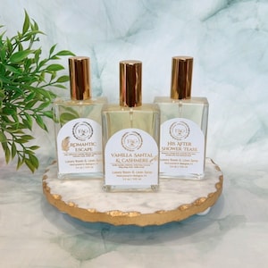 May include: Three glass spray bottles with gold caps and labels. The labels are white with gold lettering and feature the text "FAG", "Romantic Escape", "Vanilla Santal & Cashmere", and "His After Shower Tease". The bottles are on a white and gold marble-like surface.