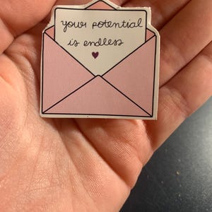 Könnte beinhalten: Rosaer Briefumschlag-Aufkleber mit einem weißen Zettel im Inneren, auf dem "your potential is endless" mit einem kleinen lila Herz steht.