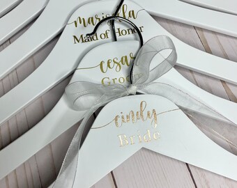 May include: Set of white wooden hangers with gold lettering. The hangers are personalized with names and titles, including "Bride", "Maid of Honor", "Best Man", and "Groom".
