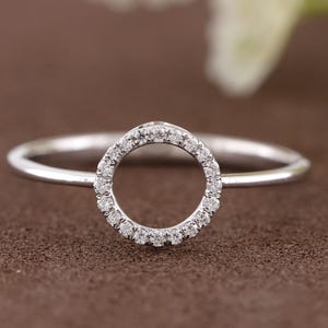 Puede incluir: Un anillo de plata con un dise&ntilde;o circular incrustado con peque&ntilde;as piedras preciosas transparentes. La banda del anillo es delgada y pulida, y las piedras preciosas est&aacute;n engastadas alrededor del c&iacute;rculo. El anillo se muestra sobre una superficie marr&oacute;n.