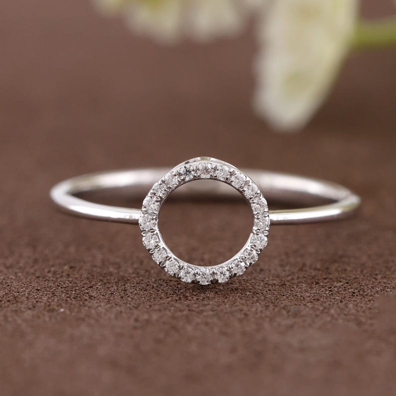 Puede incluir: Un anillo de plata con un dise&ntilde;o circular incrustado con peque&ntilde;as piedras preciosas transparentes. La banda del anillo es delgada y pulida, y las piedras preciosas est&aacute;n engastadas alrededor del c&iacute;rculo. El anillo se muestra sobre una superficie marr&oacute;n.