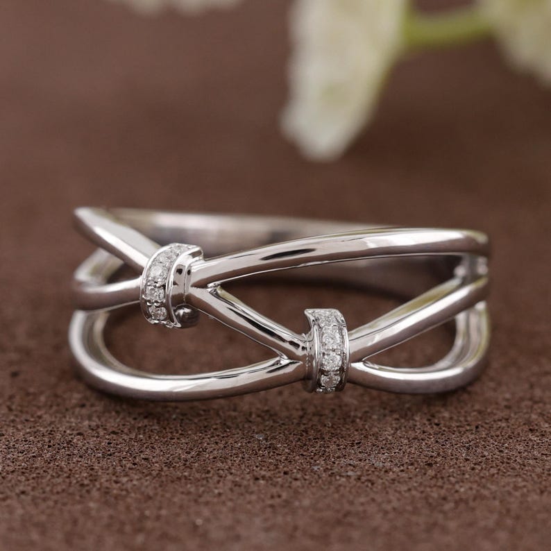 May include: A silver-colored ring with a unique, intertwined design. The ring features two small, diamond-studded accents. The band has a polished finish, creating a modern and elegant look. The ring is a piece of jewelry.