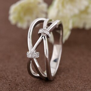 May include: A silver ring with a knot design. The ring features two bands that cross over each other, with a small section of clear stones at the knot. The ring is set against a brown background.
