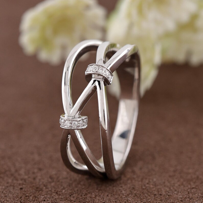 May include: A silver ring with a knot design. The ring features two bands that cross over each other, with a small section of clear stones at the knot. The ring is set against a brown background.