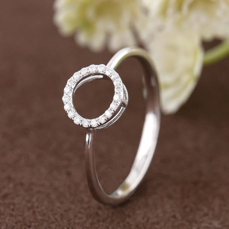 Puede incluir: Un anillo plateado con un dise&ntilde;o circular incrustado con peque&ntilde;as piedras preciosas transparentes. La banda del anillo es lisa y pulida, y las piedras preciosas est&aacute;n muy juntas, creando un efecto brillante. El anillo est&aacute; sobre un fondo marr&oacute;n suave.