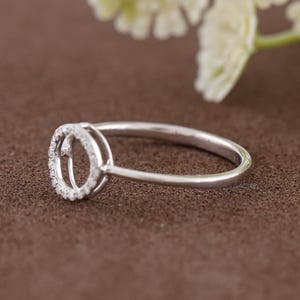 Puede incluir: Un anillo plateado con un engaste circular, incrustado con peque&ntilde;as piedras preciosas transparentes. La banda del anillo es fina y pulida, y el engaste es de dise&ntilde;o halo. El anillo est&aacute; sobre un fondo marr&oacute;n, con una flor blanca borrosa en el fondo.