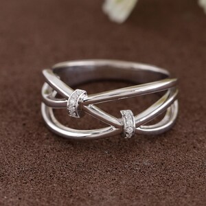 May include: A silver ring with a unique design. The ring features a series of intertwined bands, with two sections adorned with small, sparkling stones. The ring is set against a brown background.
