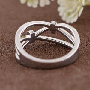 May include: A silver ring with a unique design, featuring two small square-shaped gemstones. The ring has an intertwined, openwork design. The ring is set against a brown background, with a blurred white flower in the background.