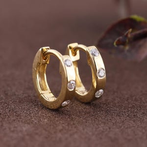 May include: A pair of small gold hoop earrings featuring a hinged closure mechanism. Each hoop is accented with three evenly spaced, round, clear, sparkling stones embedded along the outer surface of the polished gold metal band.