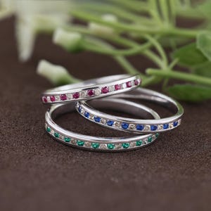 May include: Three thin silver-toned metal rings stacked together. Each ring features a channel setting with a continuous row of small, round, faceted gemstones. The top ring has red stones, the middle ring has blue stones, and the bottom ring has green stones.