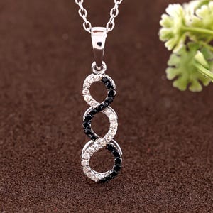 May include: A silver pendant necklace featuring an infinity symbol design. The pendant is composed of three interconnected figure-eight shapes, alternating between black and white gemstones. The necklace hangs on a delicate silver chain.