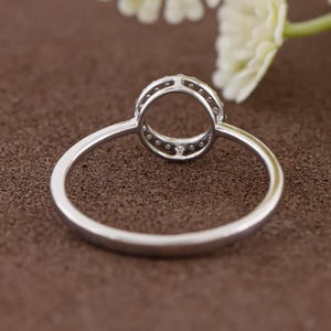 Puede incluir: Un anillo plateado con un dise&ntilde;o circular, con peque&ntilde;as piedras transparentes alrededor del borde. El anillo se muestra sobre un fondo marr&oacute;n, con una flor blanca borrosa en la esquina superior derecha. La banda del anillo es delgada y pulida.