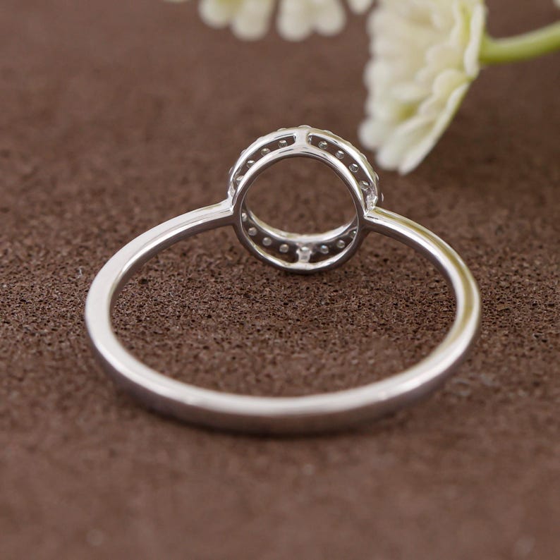 Puede incluir: Un anillo plateado con un dise&ntilde;o circular, con peque&ntilde;as piedras transparentes alrededor del borde. El anillo se muestra sobre un fondo marr&oacute;n, con una flor blanca borrosa en la esquina superior derecha. La banda del anillo es delgada y pulida.